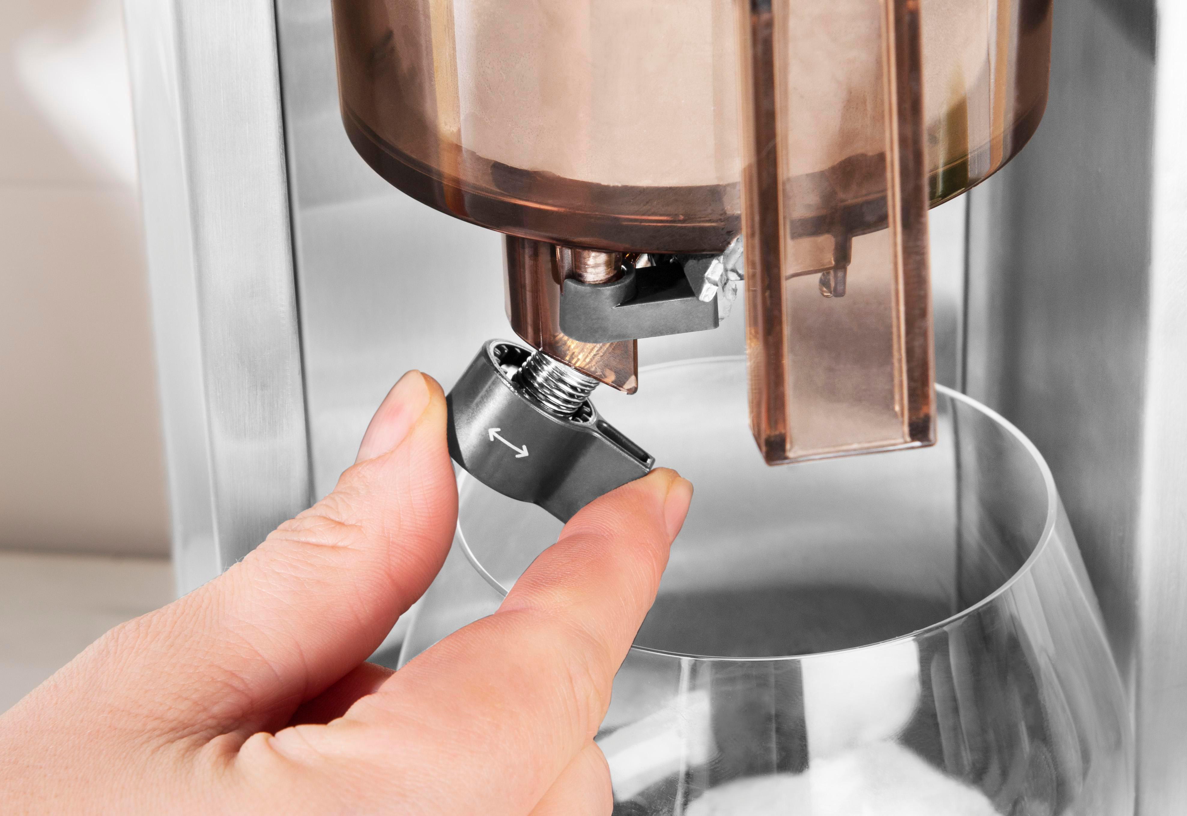 Hand verstellt einen schwarzen Hebel an einer Kaffeemaschine. Die Maschine hat einen kupferfarbenen Behälter und ein Glas darunter.