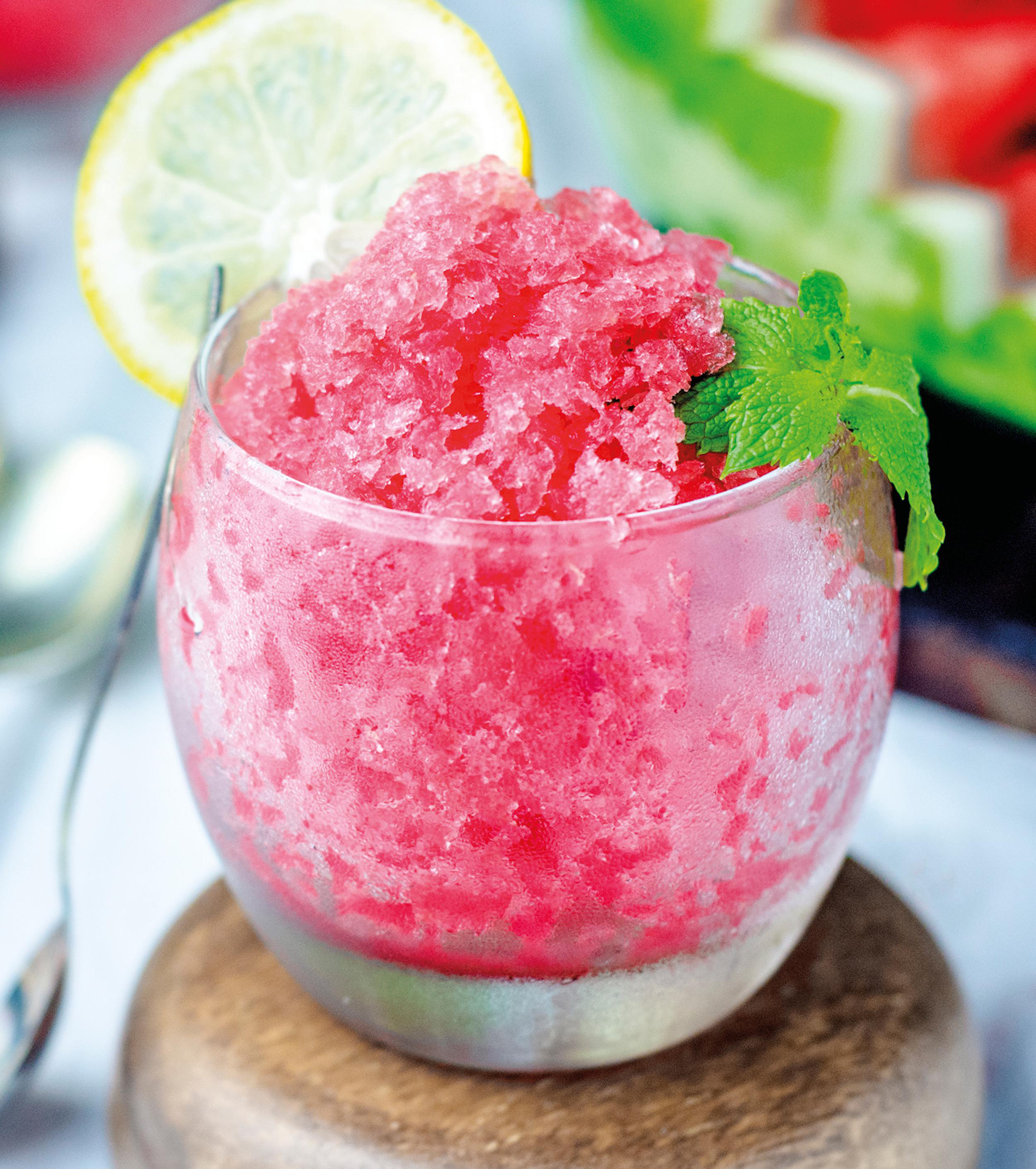 Ein Glas rosa Wassermelonen-Granita mit Zitronen- und Minzdekor, auf einem Holzuntersetzer, mit einem Löffel.
