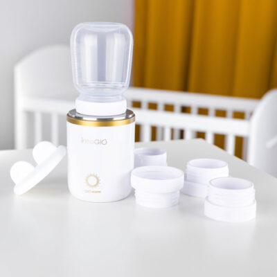 Biały i złoty podgrzewacz do butelek dla niemowląt z częściami na białym stole. W tle kołyska i żółte zasłony.