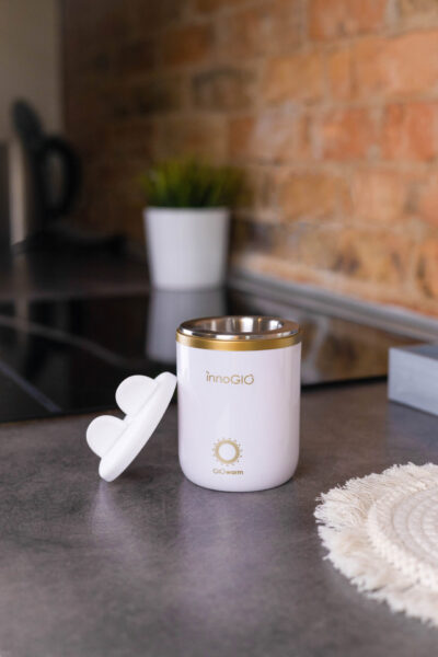 Biała podgrzewacz do butelek dla niemowląt z pokrywką stoi na blacie. Roślina w białej doniczce i ceglana ściana są w tle.