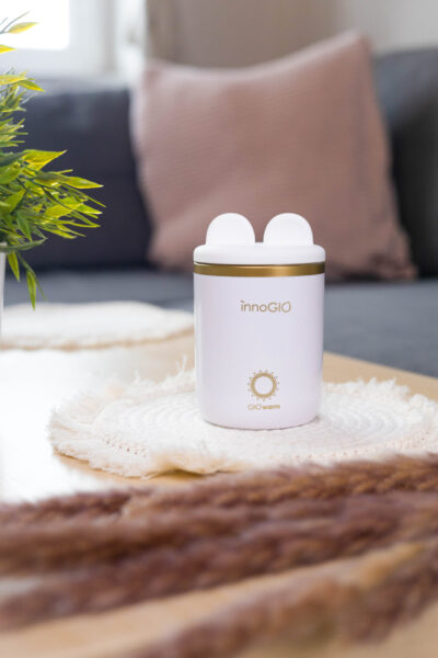 Biały podgrzewacz do butelek dla niemowląt ze złotym wykończeniem, na białej serwetce. Sofa i roślina w tle, brązowa trawa na pierwszym planie.