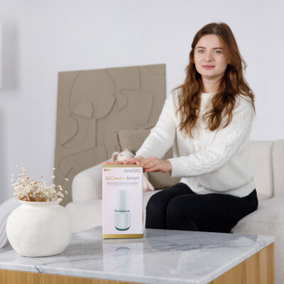 Kobieta siedzi trzymając pudełko z napisem 'GIOwarm Smart'. Ma na sobie biały sweter i czarne spodnie. W wazonie na pierwszym planie są kwiaty.