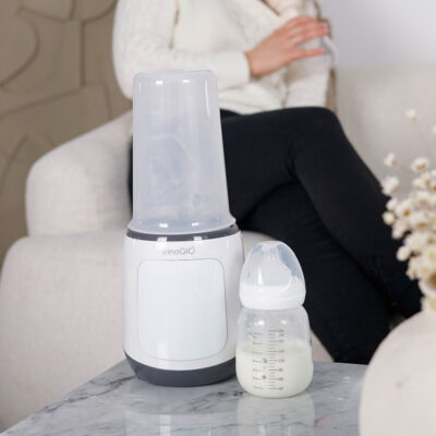 Podgrzewacz do butelek i butelka z mlekiem stoją na stole. Osoba siedzi na kanapie w tle.