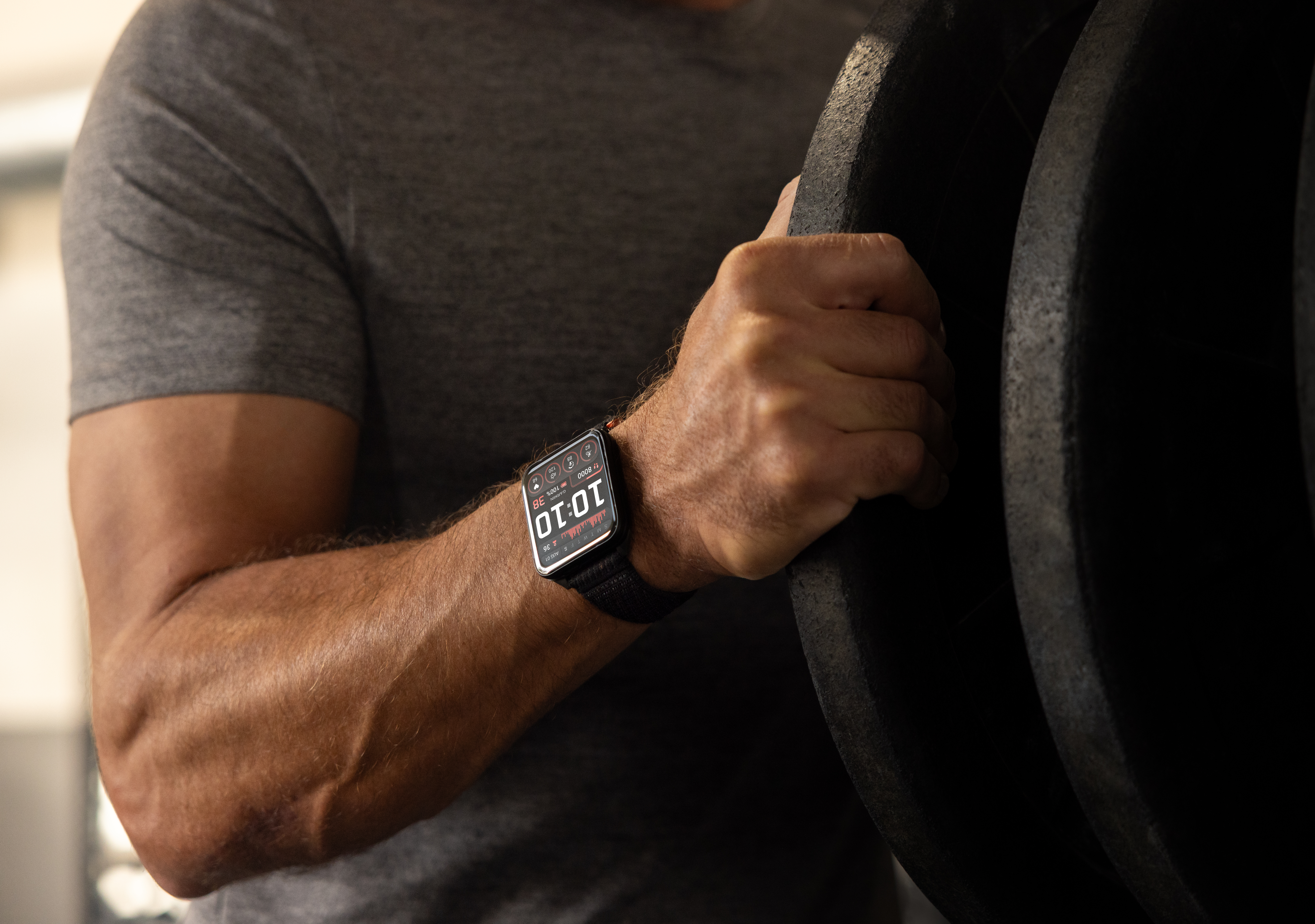 Homme avec une smartwatch tenant des poids dans une salle de sport. La montre affiche 10h10. Bras et chemise de l'homme visibles.