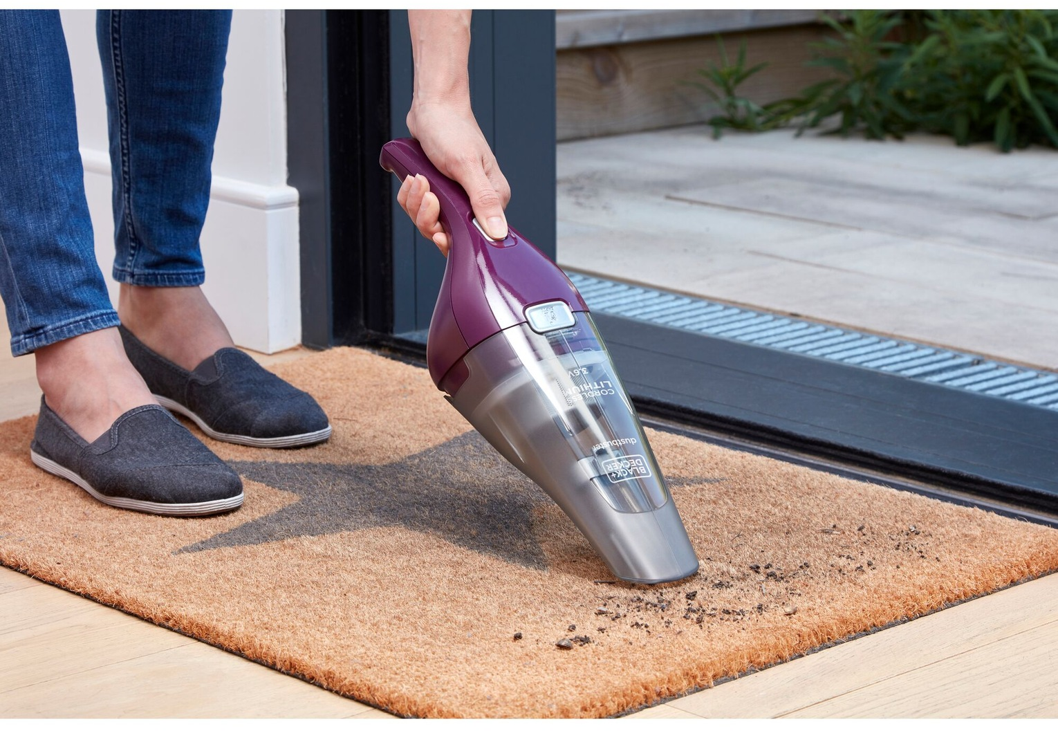 Eine Person saugt eine Fußmatte mit einem Handstaubsauger. Der Staubsauger ist lila und grau. Person trägt Jeans und graue Schuhe.