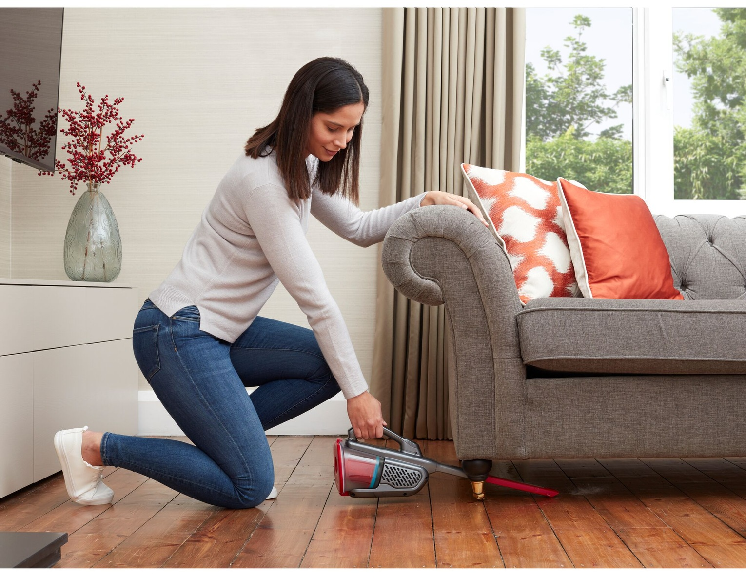 Eine Frau saugt unter einem Sofa. Sie kniet auf einem Holzboden mit einem Handstaubsauger. Links steht eine Vase mit roten Zweigen.