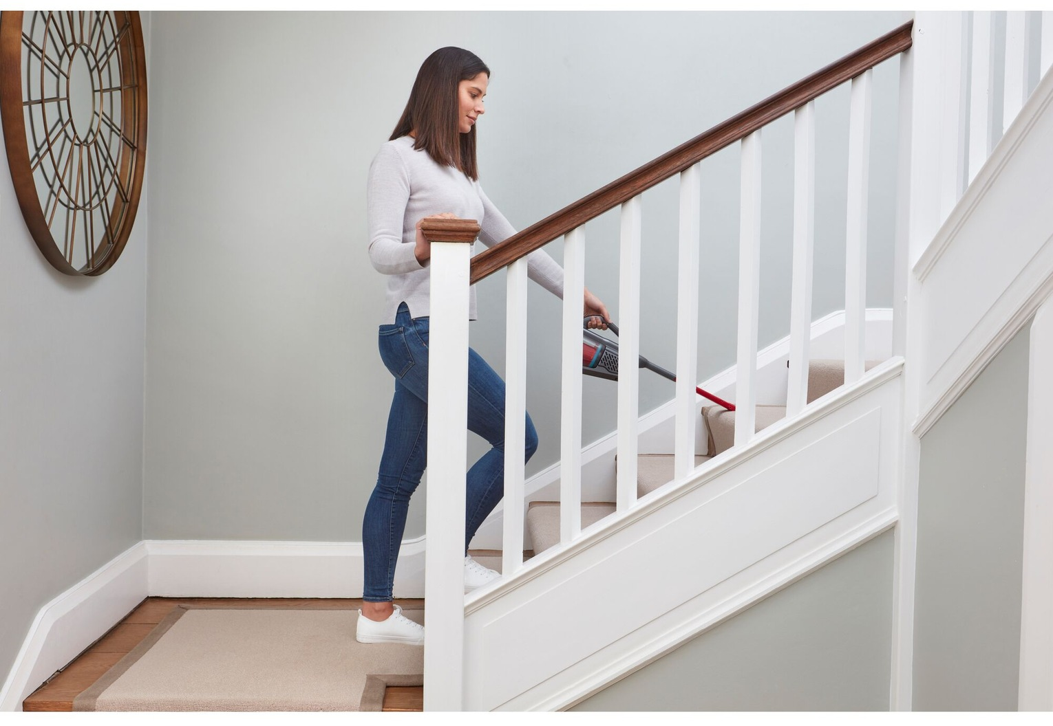 Eine Frau saugt Treppen mit einem Handstaubsauger. Sie trägt Jeans und einen Pullover. Die Treppe hat ein weißes Geländer.