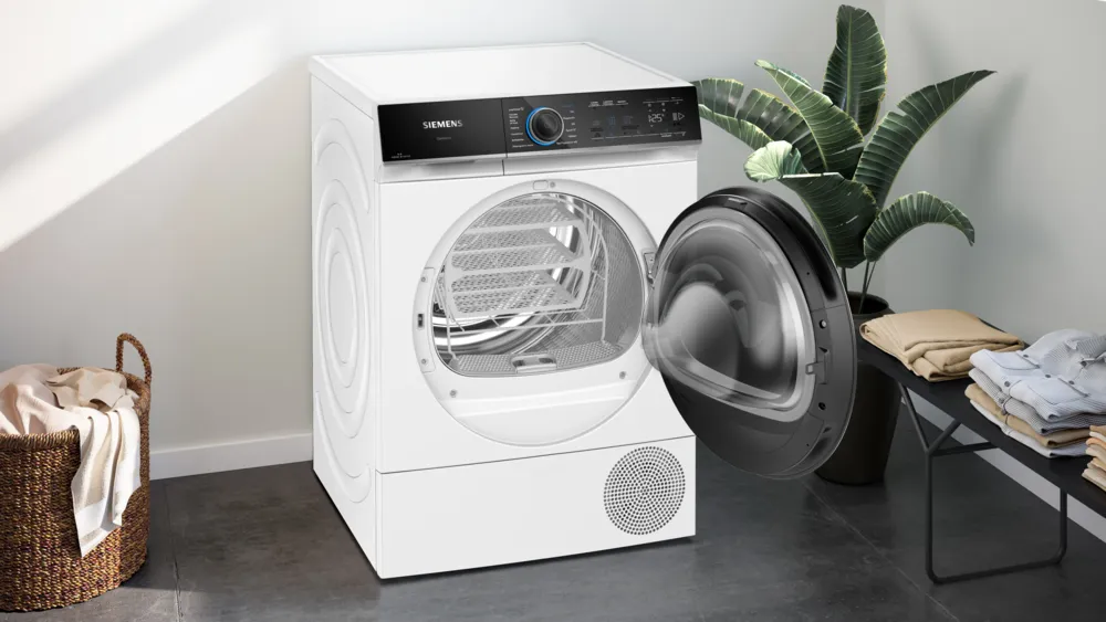 Ein weißer Siemens-Trockner mit offener Tür, Wäschekorb, Pflanze und Kleidung auf einem Tisch.