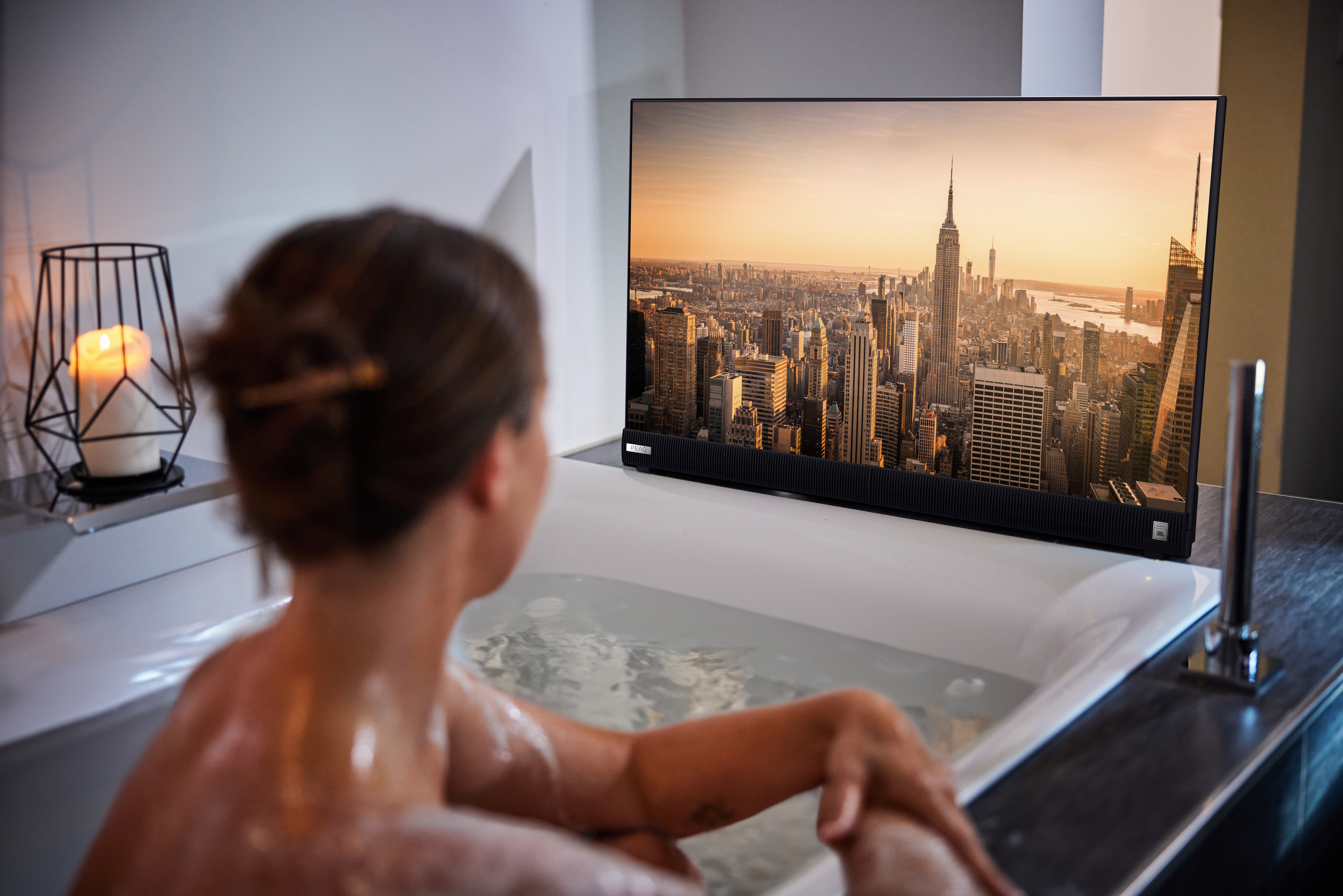 Eine Frau in der Badewanne sieht fern. Auf dem TV ist eine Stadt zu sehen. Eine Kerze steht daneben.