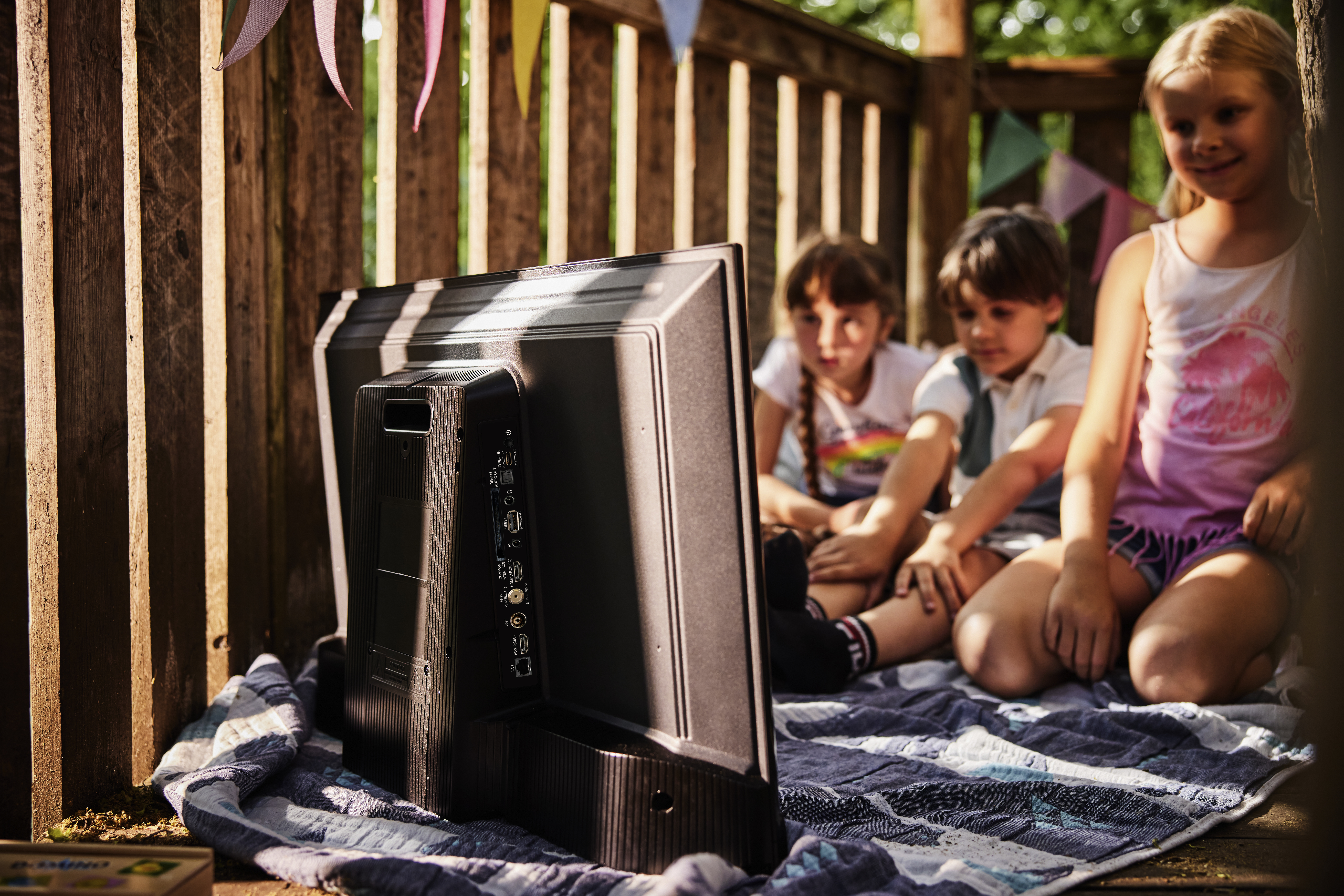 Vier Kinder in einer Holzkonstruktion schauen fern. Die Rückseite des Fernsehers ist sichtbar. Wimpel hängen über ihnen.