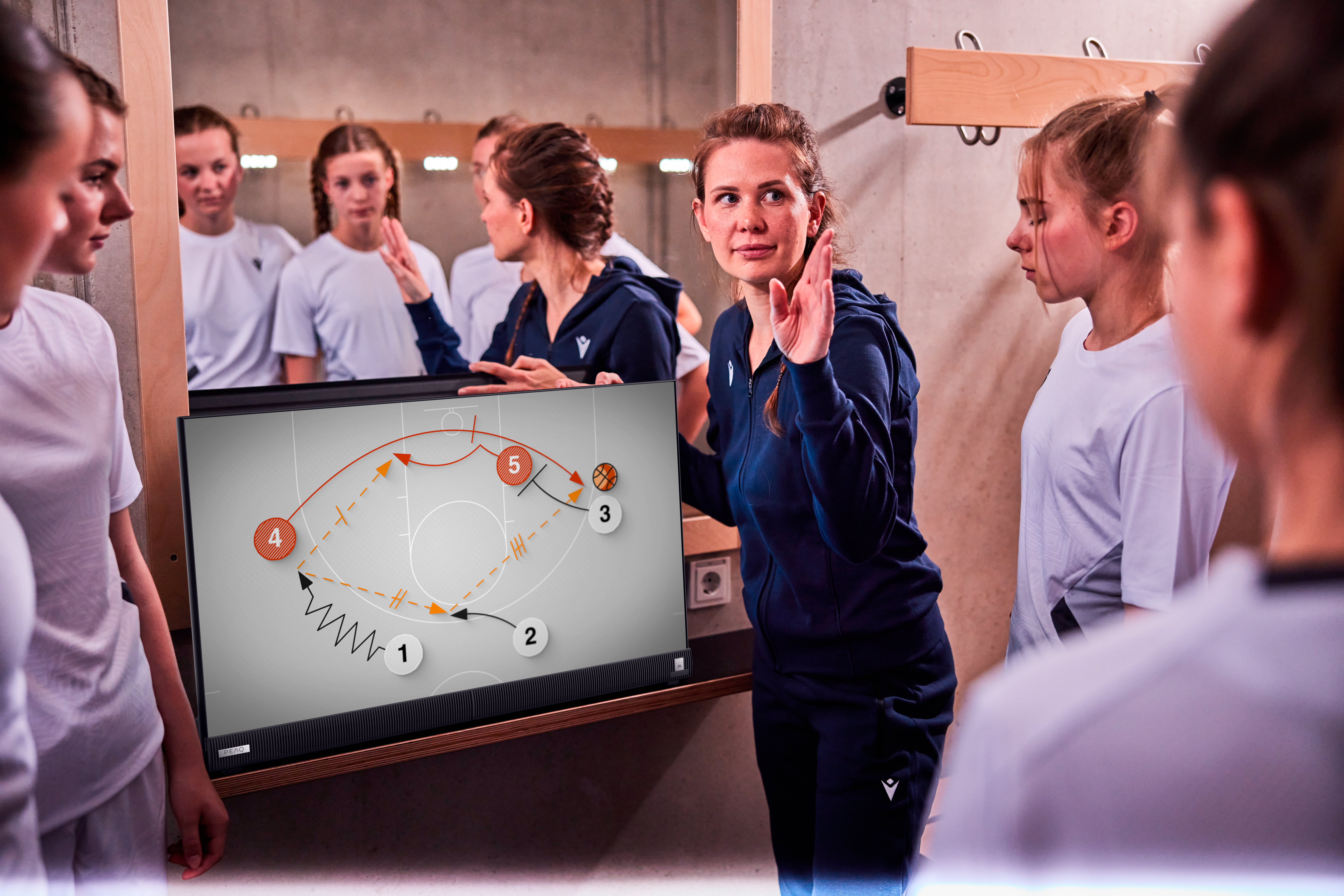Eine Frau erklärt einer Gruppe von Mädchen ein Basketballspiel. Ein Diagramm ist auf einem Bildschirm. Der Hintergrund ist eine Umkleidekabine.