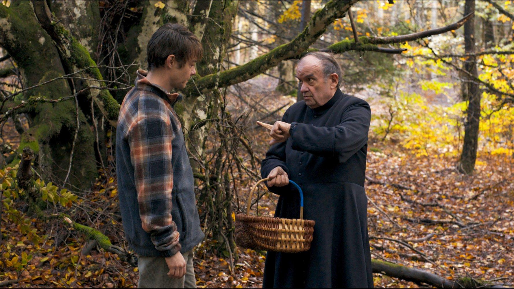 Ein Mann in einer karierten Jacke steht vor einem Priester. Der Priester zeigt mit dem Finger und hält einen Korb in einem Wald.