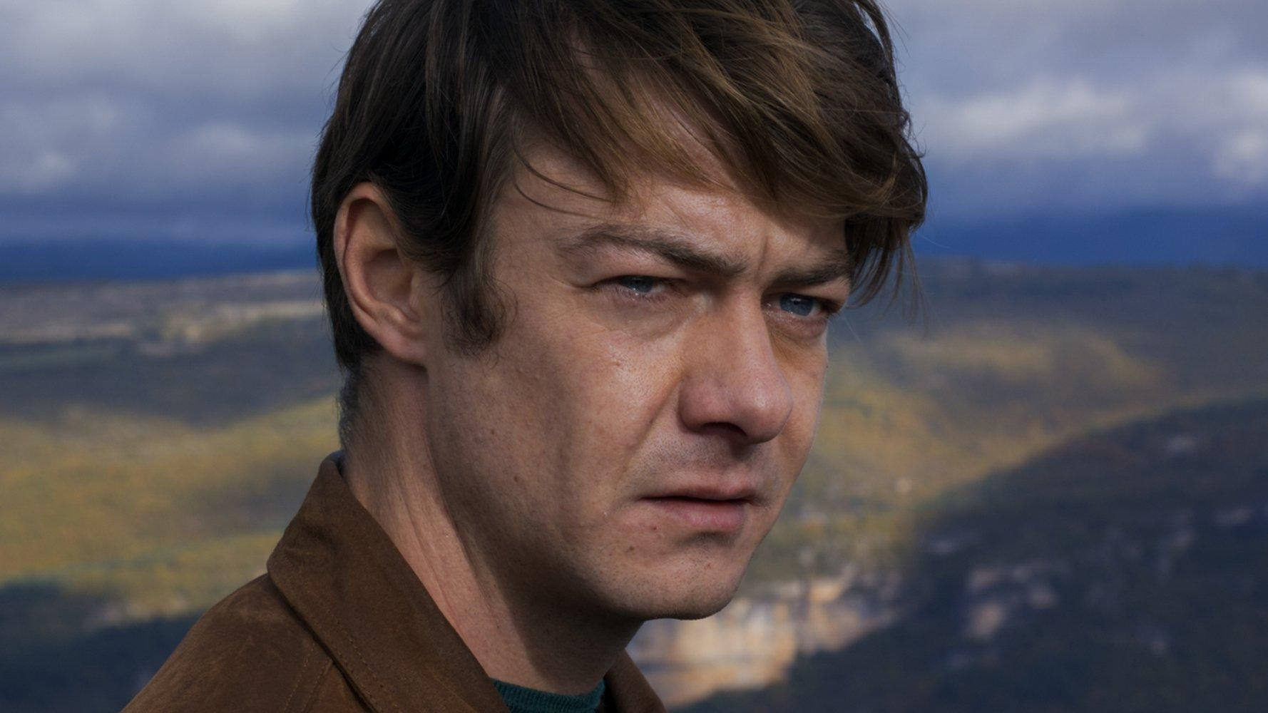Ein Mann mit kurzen braunen Haaren und blauen Augen, mit einem braunen Jackett. Verschwommene Landschaft im Hintergrund.