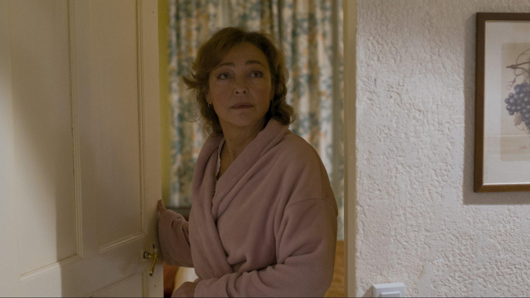 Eine Frau in einem rosa Bademantel steht in einer Tür. Sie schaut zur Seite. Ein Bild von Trauben hängt an der Wand.