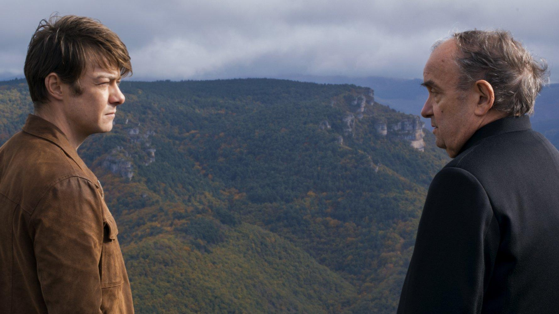 Zwei Männer blicken sich an, Berge im Hintergrund. Einer trägt eine braune Jacke, der andere einen schwarzen Mantel.