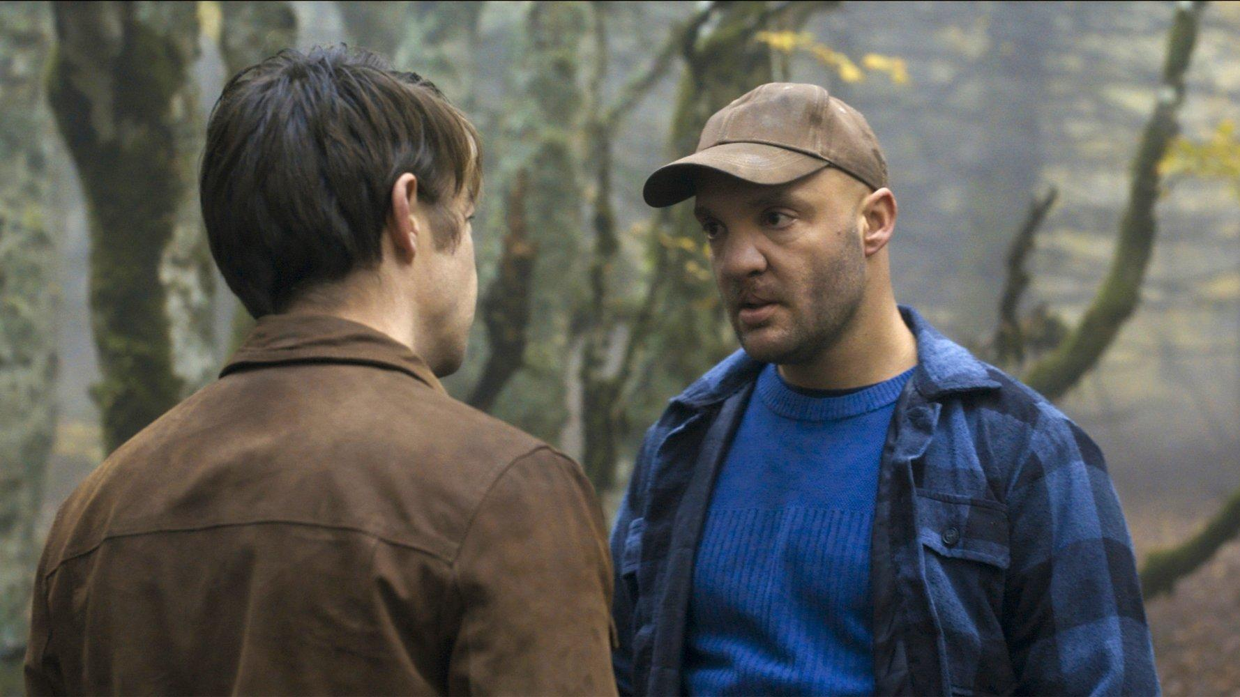 Zwei Männer im Wald. Einer trägt eine braune Mütze und ein kariertes Hemd, der andere eine braune Jacke. Bäume im Hintergrund.