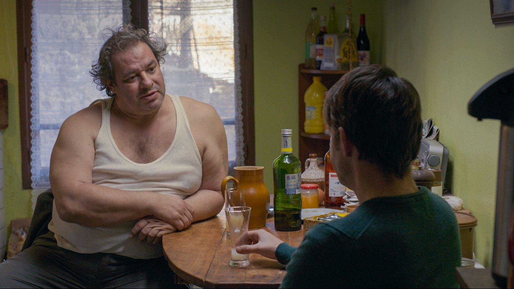 Zwei Männer sitzen an einem Tisch. Einer, im Tanktop, schaut den anderen an, der ein Glas hält. Flaschen und ein Krug stehen auf dem Tisch.