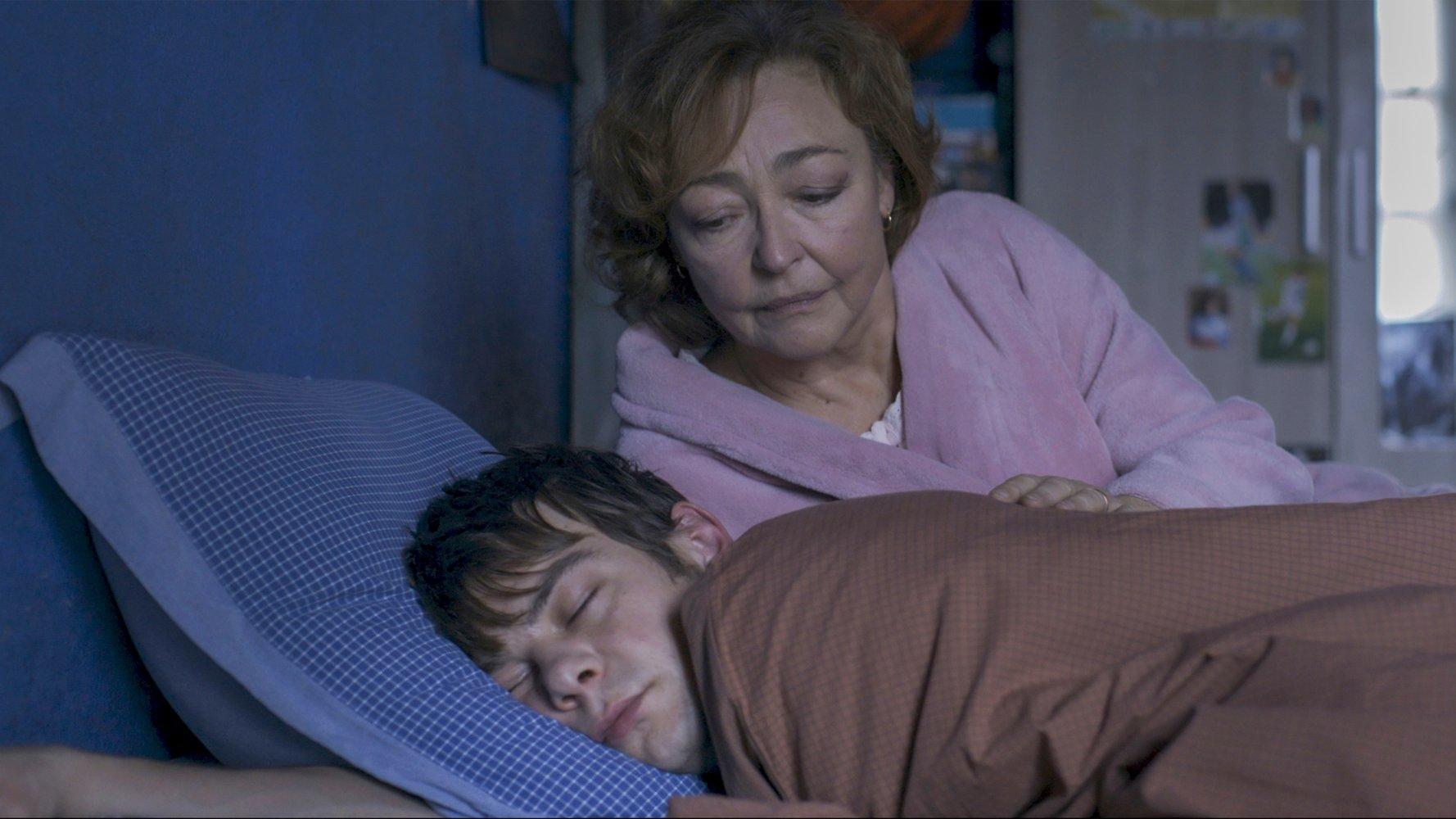 Eine Frau im rosa Bademantel blickt auf einen schlafenden jungen Mann. Er liegt auf einem blauen Kissen, bedeckt von einer braunen Decke.