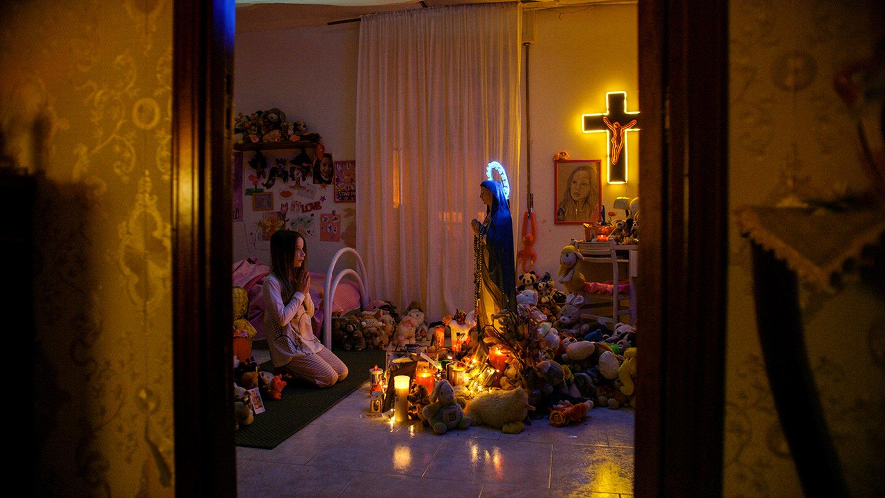 Ein betendes Mädchen in einem Raum voller Spielzeug und religiöser Symbole. Kerzen und eine Marienstatue sind vorhanden.