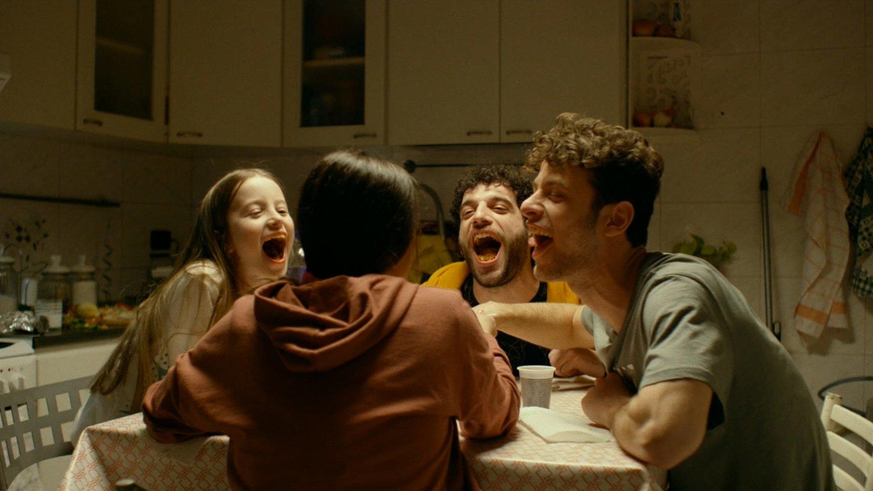 Vier Personen lachen an einem Tisch. Im Hintergrund ist eine Küche mit Schränken zu sehen. Der Tisch ist mit einer Tischdecke bedeckt.