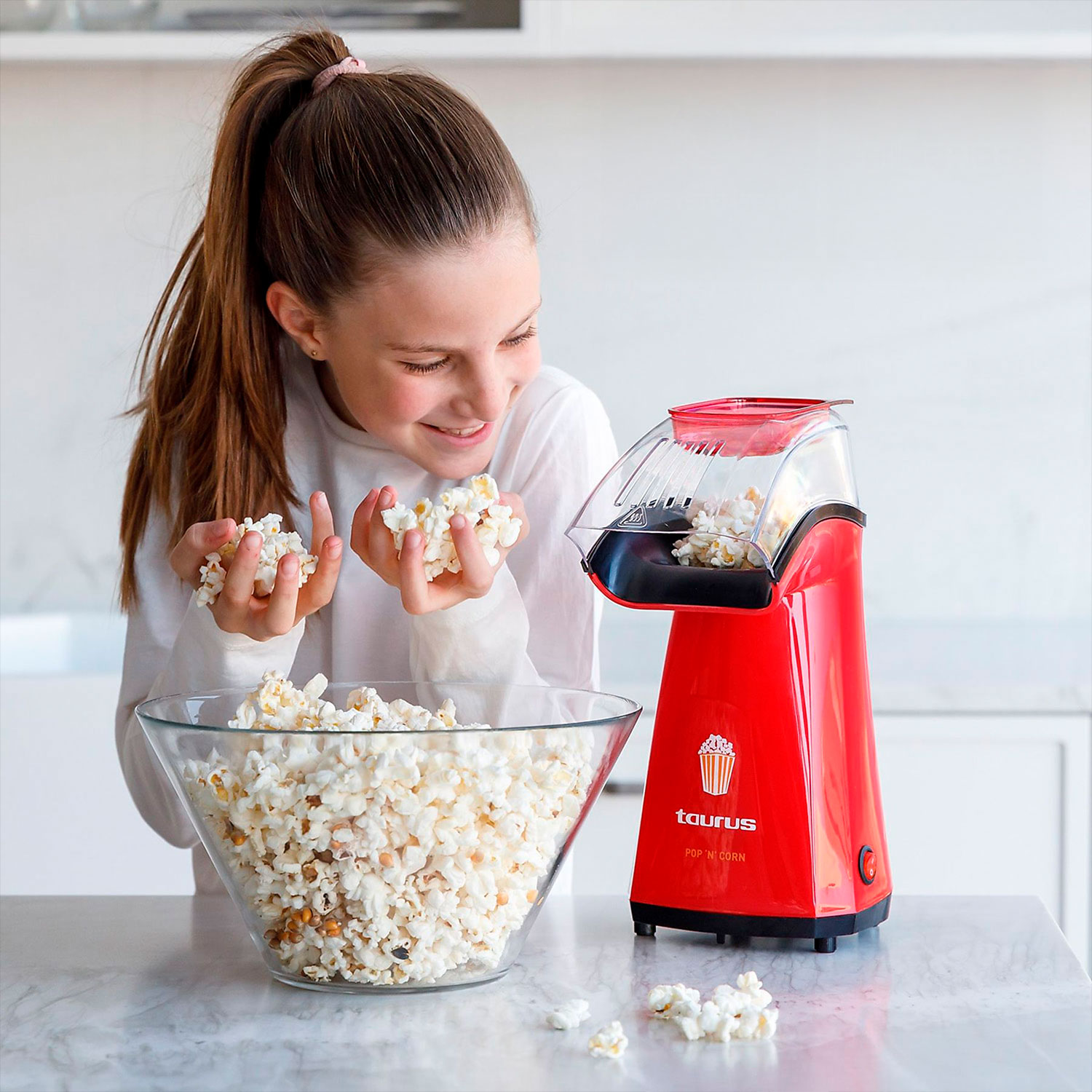 Una niña sostiene palomitas de maíz, sonriendo a una máquina roja de palomitas. Un cuenco de cristal con palomitas está junto a la máquina.