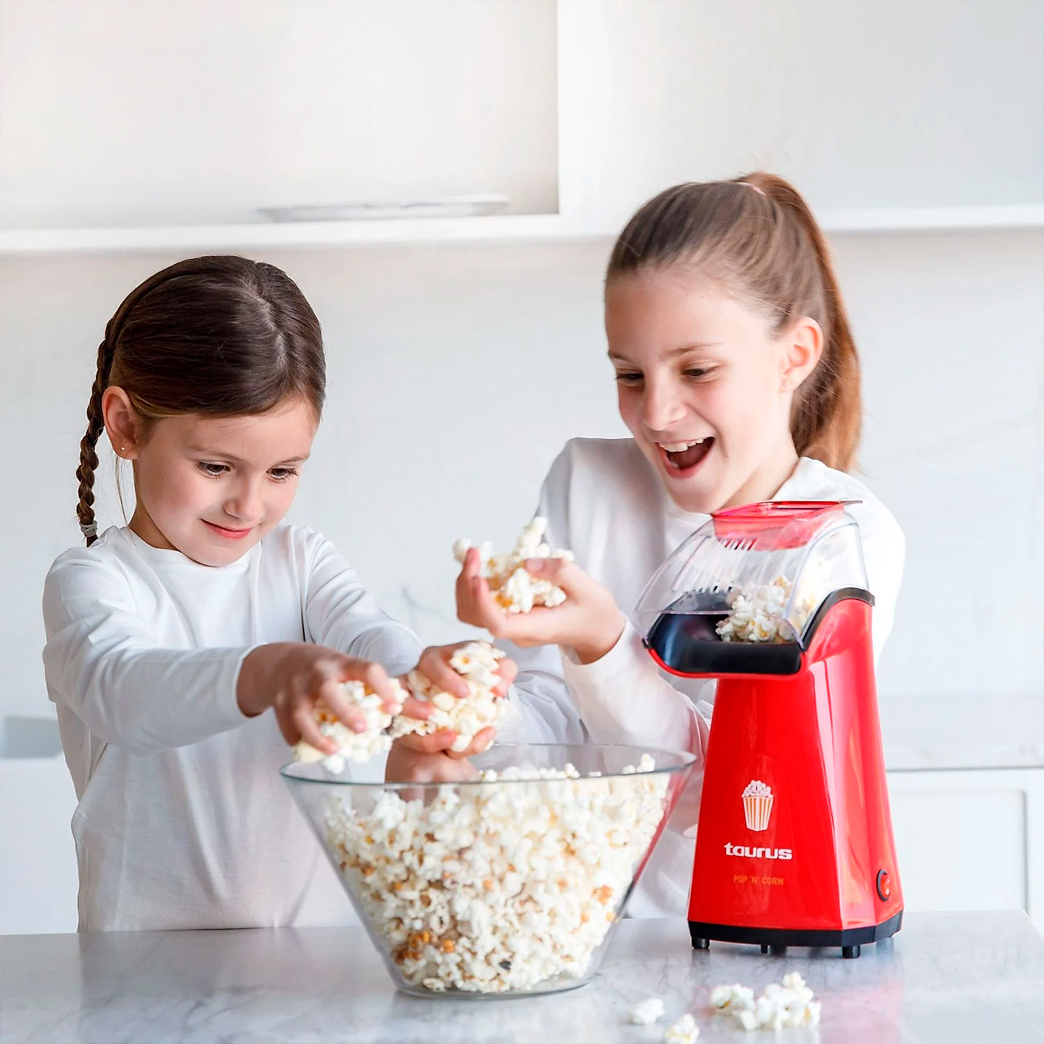 Dos niñas con una máquina de palomitas roja y un tazón de palomitas de maíz. Están sonriendo y manipulando las palomitas.