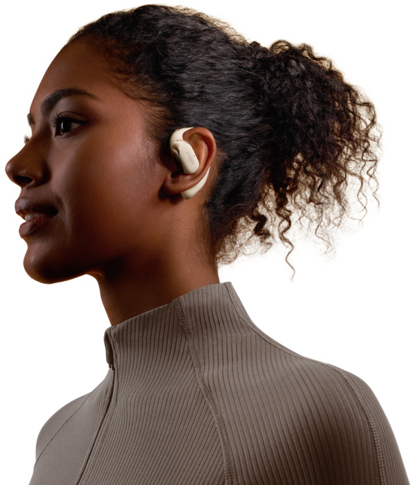 Une femme à la peau foncée porte un écouteur beige, regardant vers la gauche. Elle porte une chemise beige zippée. Ses cheveux sont bouclés.