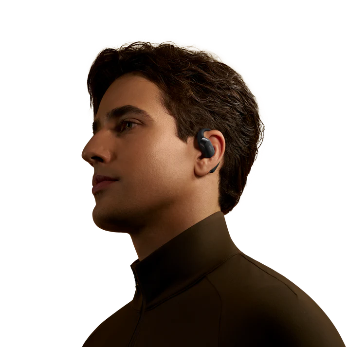 Un hombre de perfil usa un auricular en su oreja izquierda. Tiene cabello castaño y una camisa marrón oscuro. El fondo es negro.