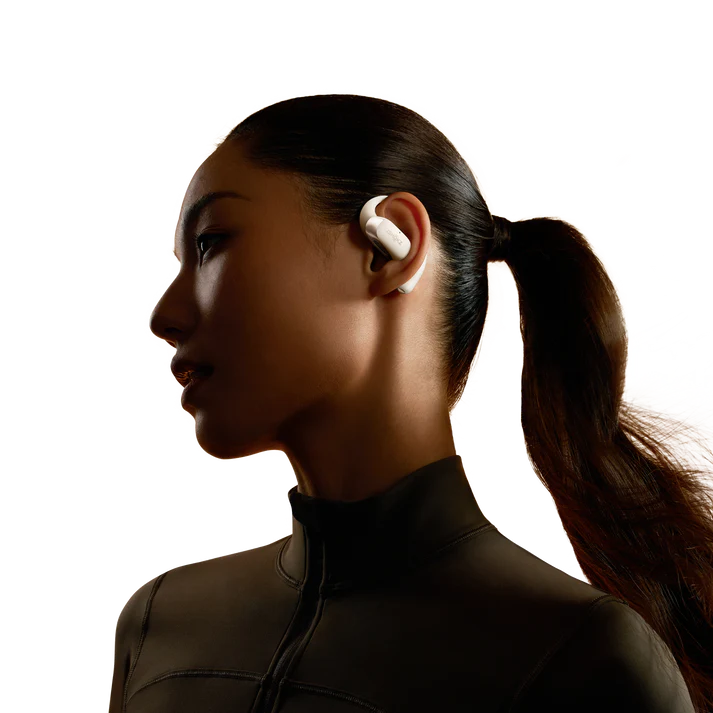 Una mujer con auriculares blancos, mirando hacia la izquierda, en un entorno oscuro. Tiene una cola de caballo y lleva una camiseta deportiva oscura.