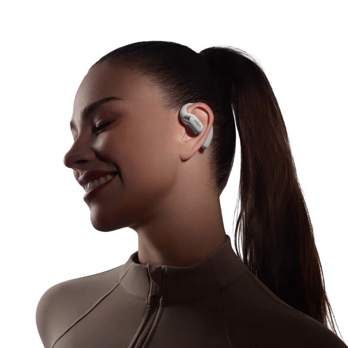Una mujer sonríe, usando un auricular gris en la oreja derecha. Su cabello está recogido en una cola de caballo. Lleva una camiseta beige.
