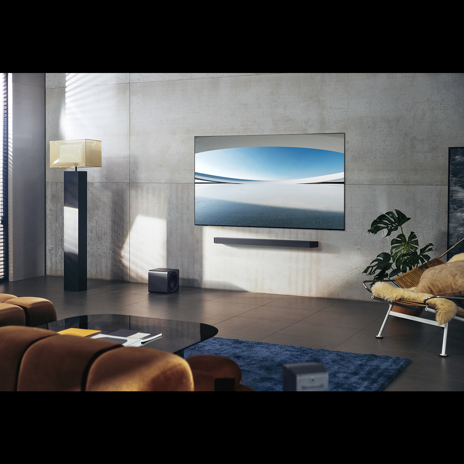 Salón moderno con TV grande, barra de sonido y subwoofer. Sofá marrón, mesa negra, alfombra azul y silla con manta.