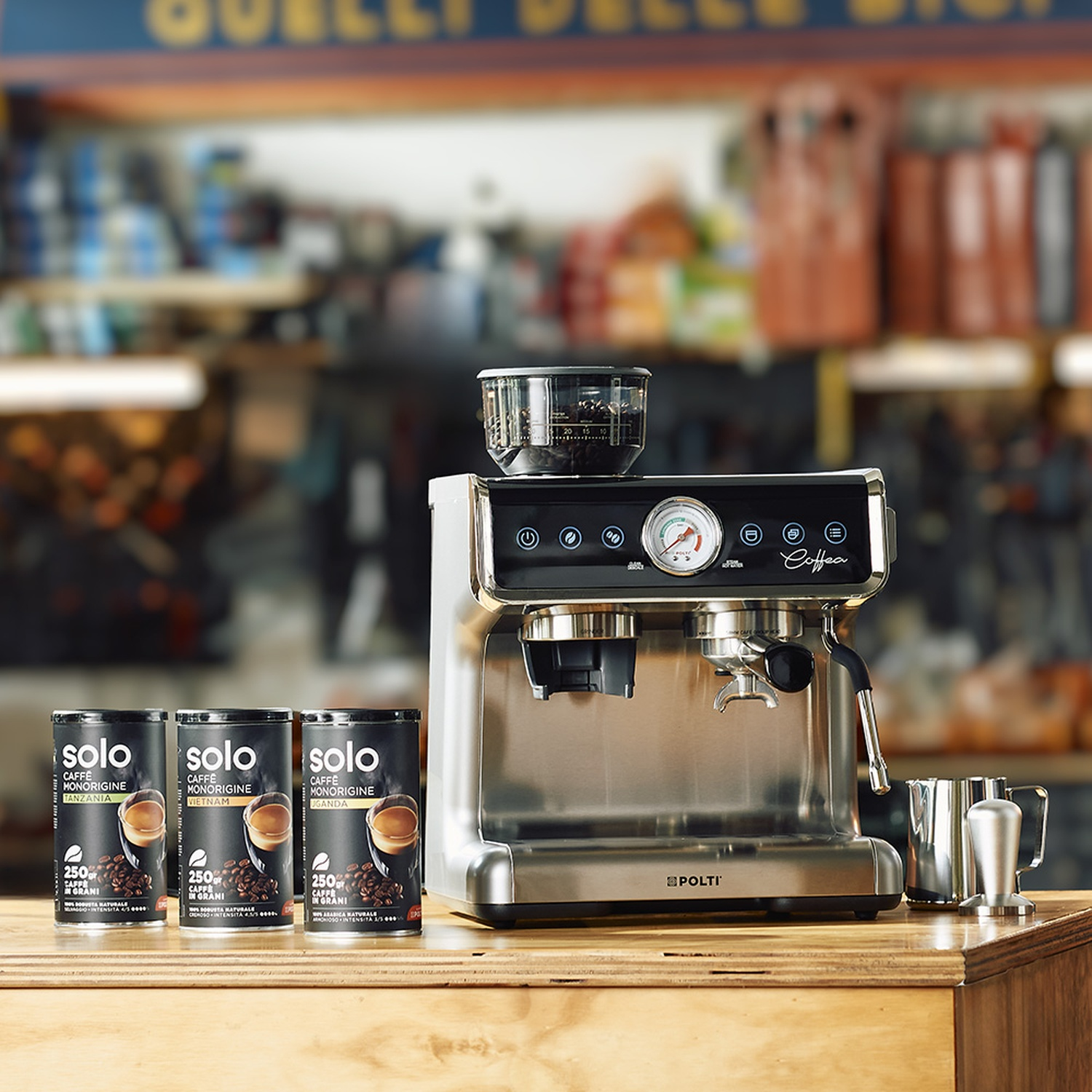 Una macchina per caffè espresso in acciaio inossidabile è appoggiata su un bancone di legno, con tre lattine di caffè e un bricco per montare il latte. Lo sfondo è sfocato.