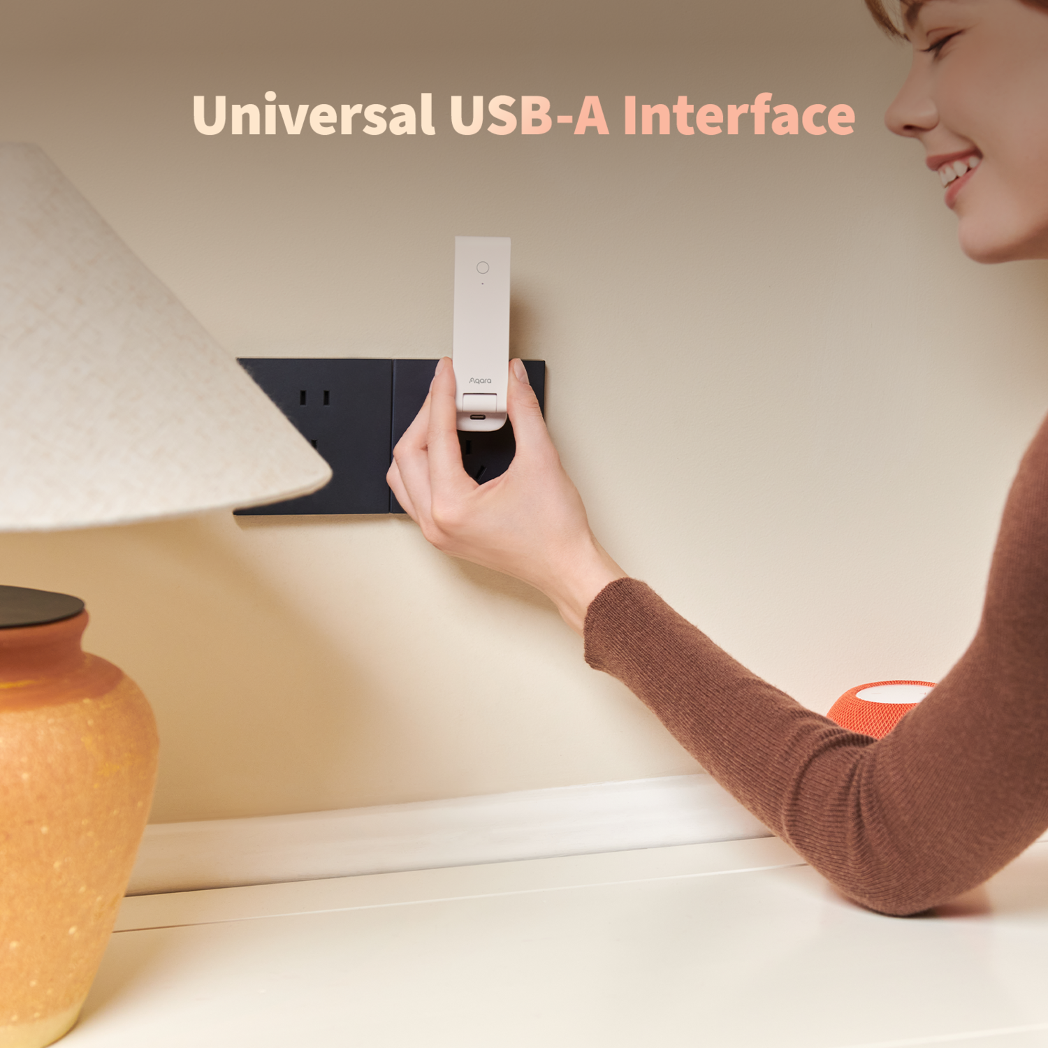A woman inserts a white USB-A adapter into a black wall outlet. A lamp and an orange speaker are also visible.