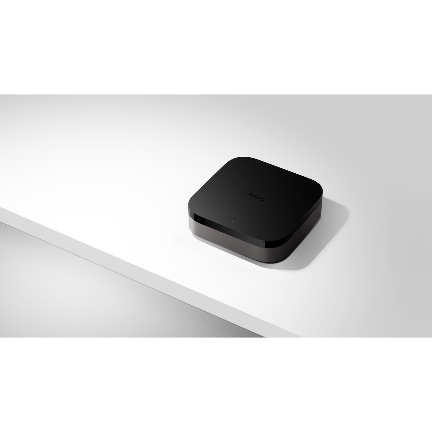A black Aqara hub sits on a white surface. The hub is square-shaped with rounded edges. The background is plain white.