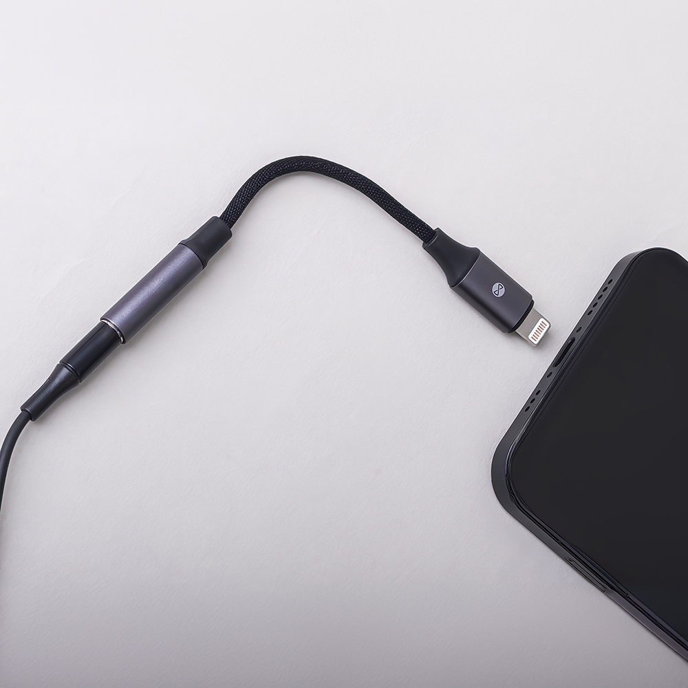 Egy fekete telefon egy töltőkábel mellett. A kábelen Lightning csatlakozó van a telefonhoz csatlakoztatva. A háttér fehér.