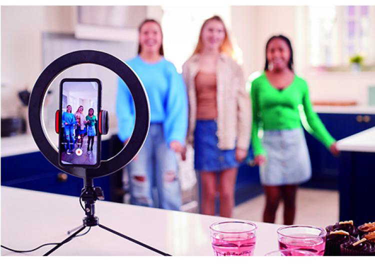Három lány videózik gyűrűs fénnyel és telefonnal. Rózsaszín italok és muffinok az előtérben. A lányok konyhában vannak.