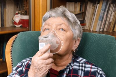 Starsza kobieta z krótkimi, siwymi włosami w masce tlenowej, siedząca na krześle. Regały z książkami w tle.