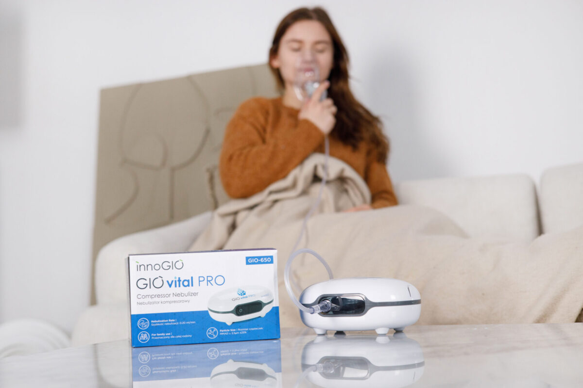 Kobieta używa nebulizatora. Nebulizator i pudełko na stole. Tło: sofa, koc i ściana.