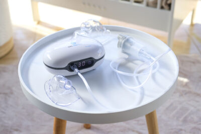 Biały nebulizator z akcesoriami stoi na małym białym stoliku. Stół ma drewniane nogi. W tle jest łóżeczko.