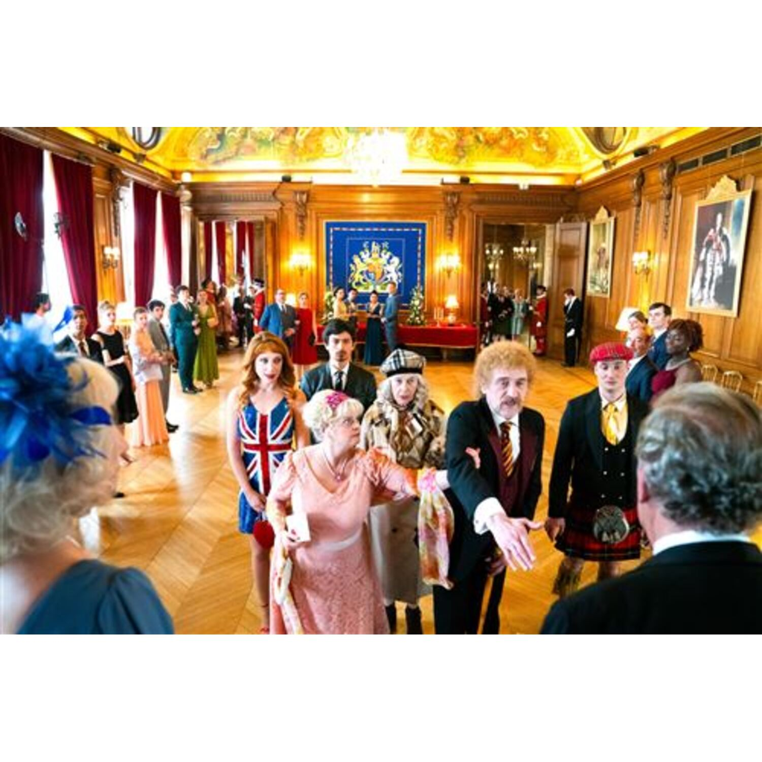 Een groep mensen in een formele zaal. Sommigen dragen formele kleding. De zaal heeft schilderijen, lambrisering en een decoratief plafond.