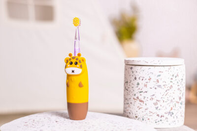Żółta szczoteczka do zębów z motywem żyrafy stoi na białej powierzchni. Po prawej stronie znajduje się ozdobny pojemnik. W tle biały namiot.