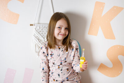 Dziewczyna uśmiecha się, trzymając żółtą szczoteczkę do zębów. Ma na sobie koszulę we wzory. Tło: biała ściana z dekoracją makramy i dużymi literami.