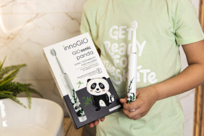 Dziecko trzyma pudełko i szczoteczkę do zębów z motywem pandy. Na pudełku napis 'GIO sonic panda', dziecko ma na sobie zieloną koszulkę.