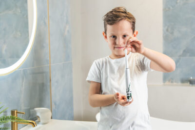 Chłopiec trzymający elektryczną szczoteczkę do zębów. W tle lustro, umywalka i niebiesko-biała kafelkowa ściana.
