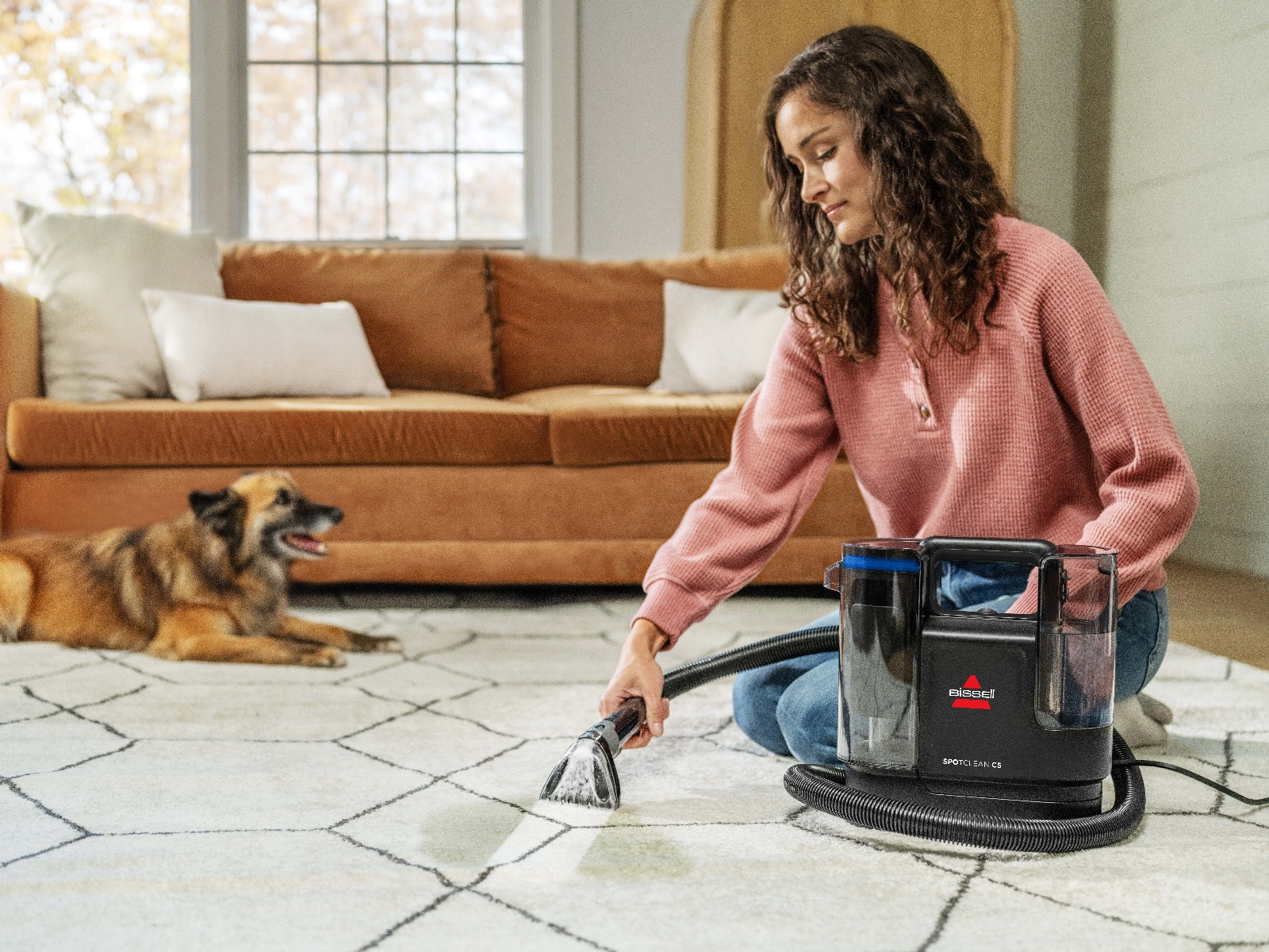 Kadın, Bissell SpotClean C5 ile halıyı temizliyor. Halının üzerinde bir köpek yatıyor. Arka planda kahverengi bir kanepe ve pencere var.