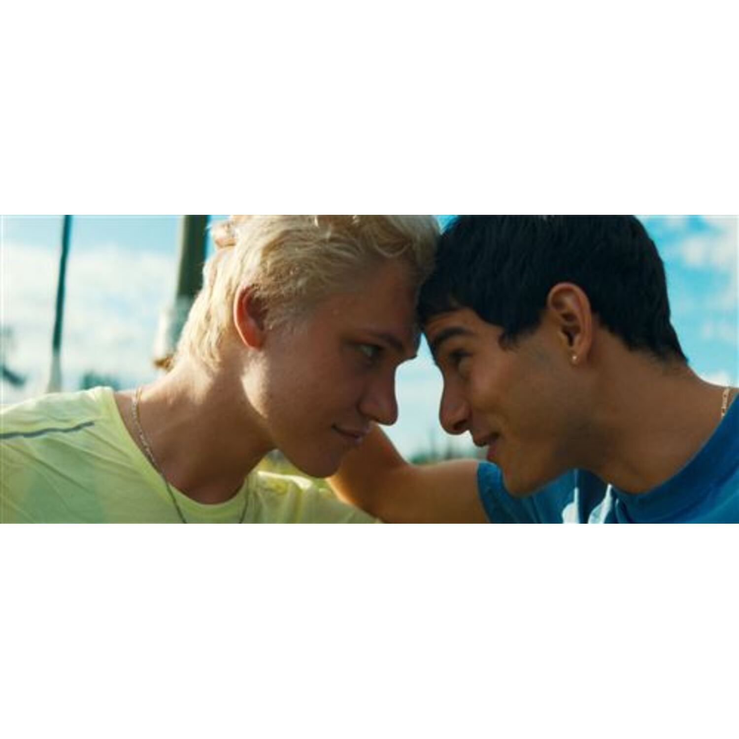 Twee jonge mannen staan tegenover elkaar, hun voorhoofden raken elkaar aan. De een heeft blond haar en een geel shirt, de ander zwart haar en een blauw shirt.