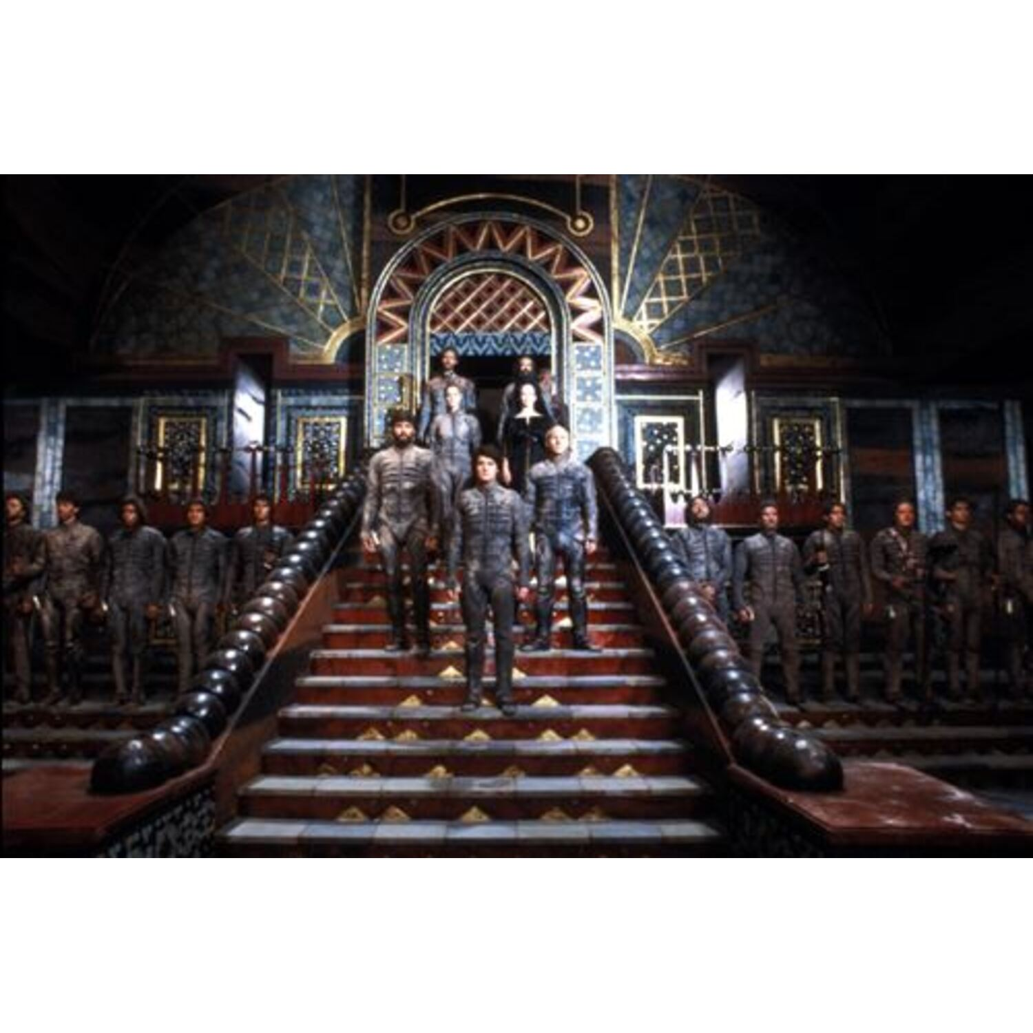 Un groupe de personnes en uniformes sombres descend un grand escalier. D'autres se tiennent sur les marches, et d'autres bordent les côtés. Décor orné.