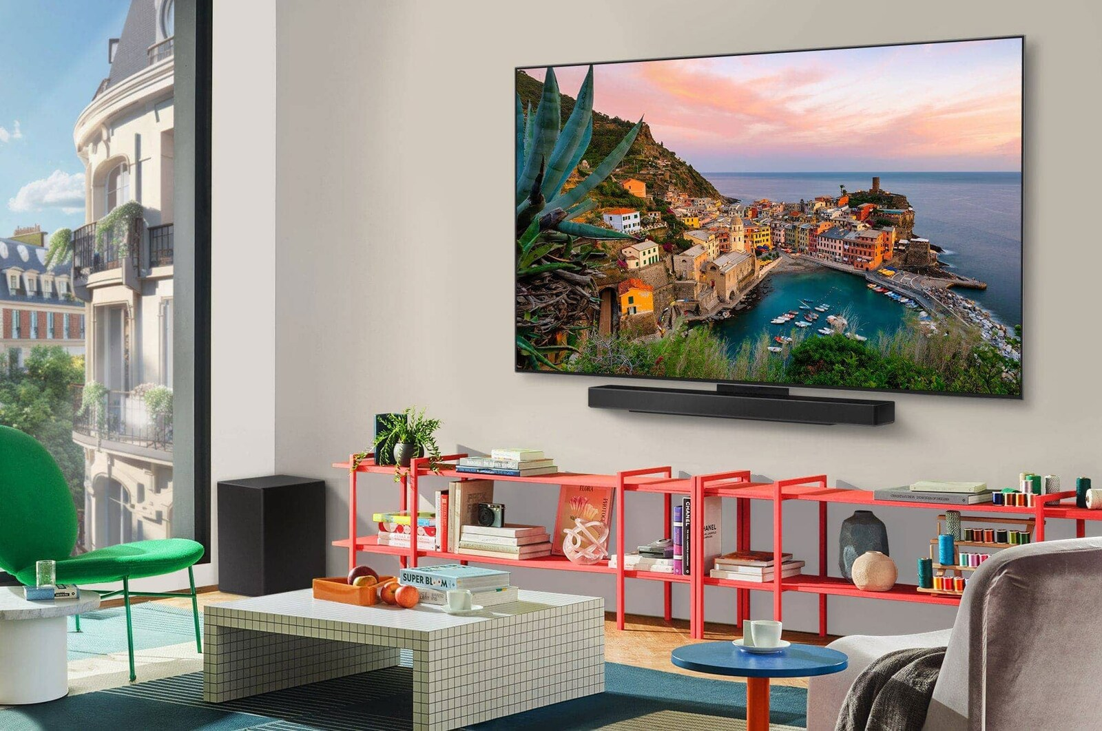 Wohnzimmer mit großem Fernseher, der eine Küstenstadt zeigt. Rote Regale mit Büchern und Dekoration. Ein grüner Stuhl und ein weißer Tisch.
