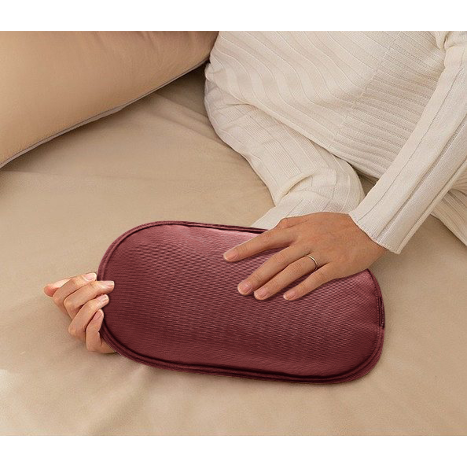Een persoon houdt een bordeauxrood kussen op een bed vast. De persoon draagt een witte trui. Het bed heeft beige lakens.