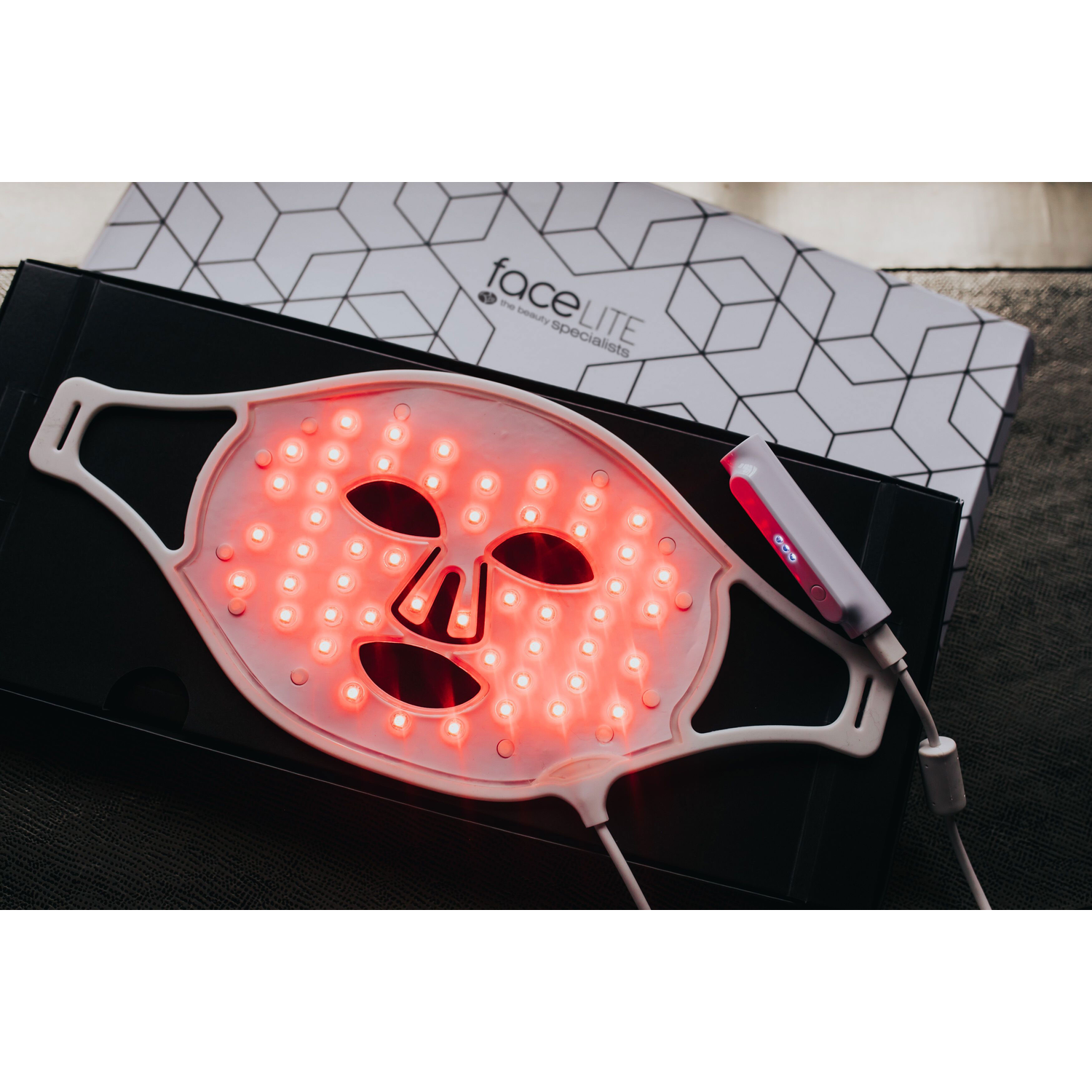 A red LED face mask, a white control device, and a box with a geometric pattern are displayed on a dark surface.